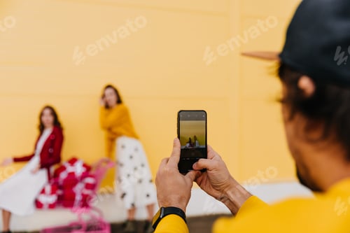 Preview: Man in black cap making photo of two girls. Cropped shot of man holding smartphone