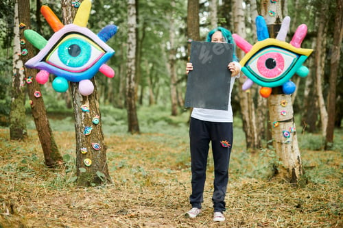 Preview: Funny young adult woman with dyed turquoise hair in white T shirt near big eye art installation