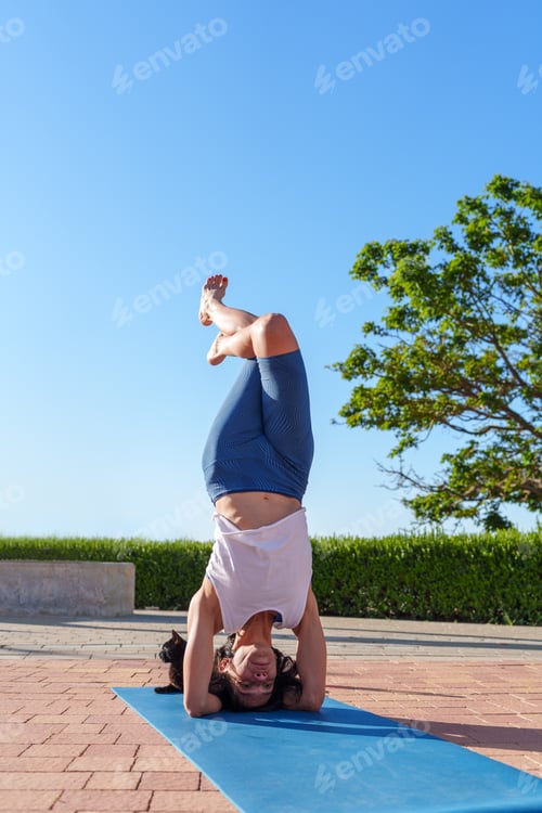 Preview: Woman performing salamba shirshasana yoga pose on mat beside resting cat, basking in sunlight