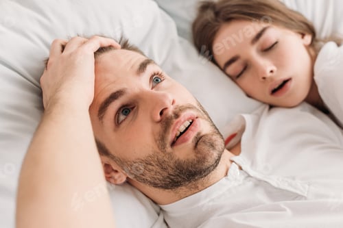 Preview: awakened man holding hand on head while lying in bed near snoring wife