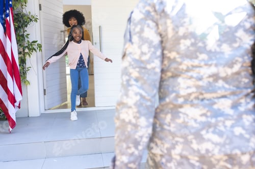 Preview: Happy african american wife and daughter welcoming male soldier outdoors with usa flag, copy space