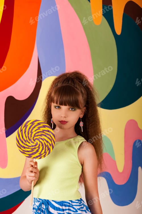 Preview: Adorable little girl holding round giant lollipop at colored wall, looking at camera