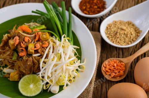 Preview: Pad Thai in a white plate with lemon, eggs and seasoning on a wooden table.