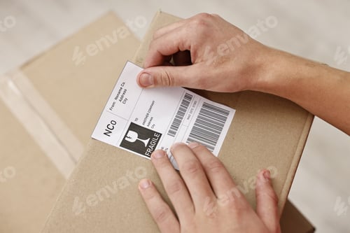 Preview: Man sticking shipping label with barcode on parcel indoors, closeup