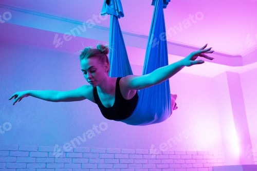 Preview: CHarming fit young woman doing fly yoga stretching with hammock in yoga studio.