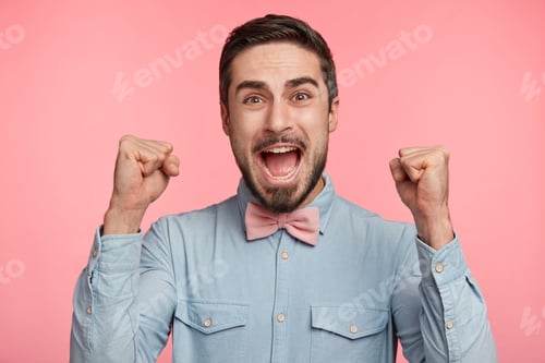 Preview: Successful European male with stubble raises fists in joy, celebrates victory or achievement goals,