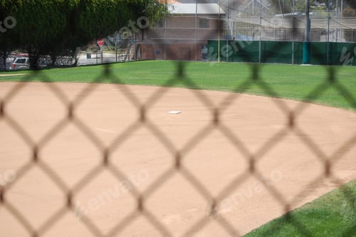 Vorschau: Verschwommener Maschendrahtzaun mit einem Baseballfeld und Gebäuden im Hintergrund unter Sonnenlicht