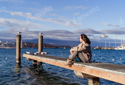 Preview: Happy young girl enjoying sunny day sitting on jetty with seagulls looking at blue lake