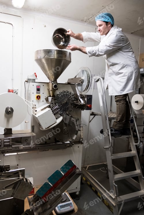 Preview: Man wearing white coat standing on a ladder, pouring tea leaves into large funnel of a machine.