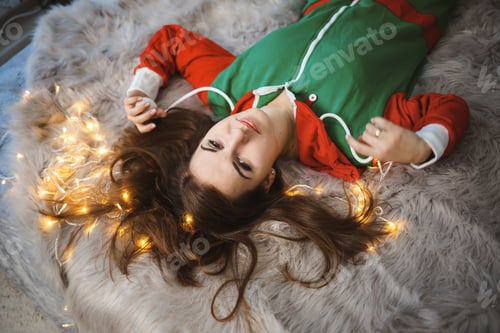 Preview: A girl in a New Year's overalls with a festive mood lies on a bed around luminous garlands