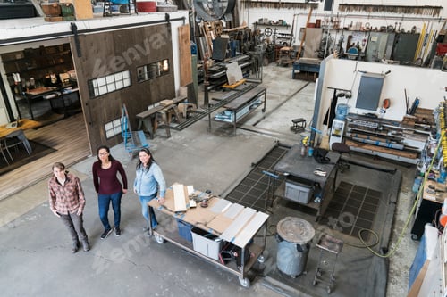 Preview: High angle view of three women standing in metal workshop, looking at camera.