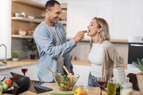 Preview: Cheerful married multinational couple using laptop while cooking healthy food in kitchen