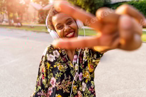 Preview: Happy fashionable young woman with headphones outdoors at sunset making victory gesture
