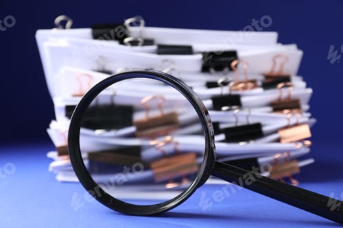 Preview: Magnifying glass and many documents with binder clips on blue background, closeup