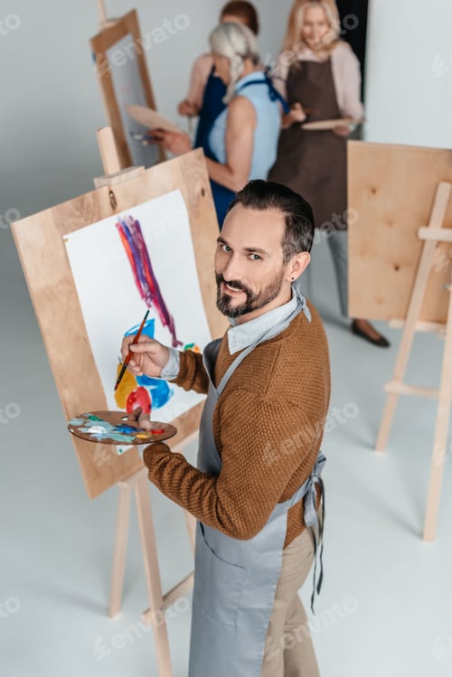 Preview: handsome mature artist holding palette while standing near easel and smiling at camera