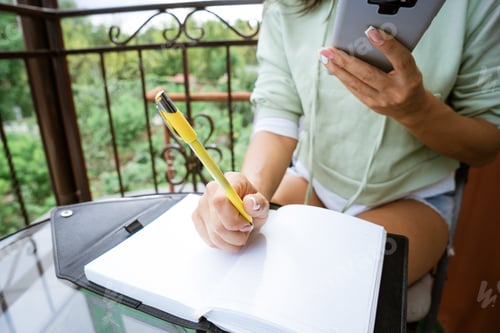 Preview: Young woman writes in notebook with phone in hand on the balcony