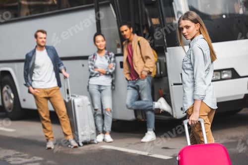 Preview: selective focus of young woman carrying wheeled bag while her friends waiting near travel bus at