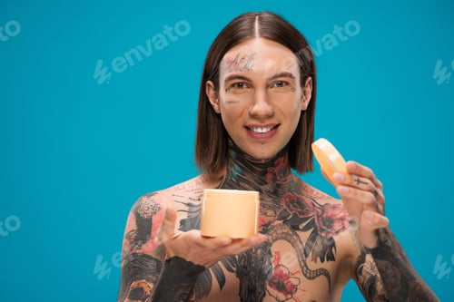 Preview: happy young man with tattoos holding container with body butter isolated on blue