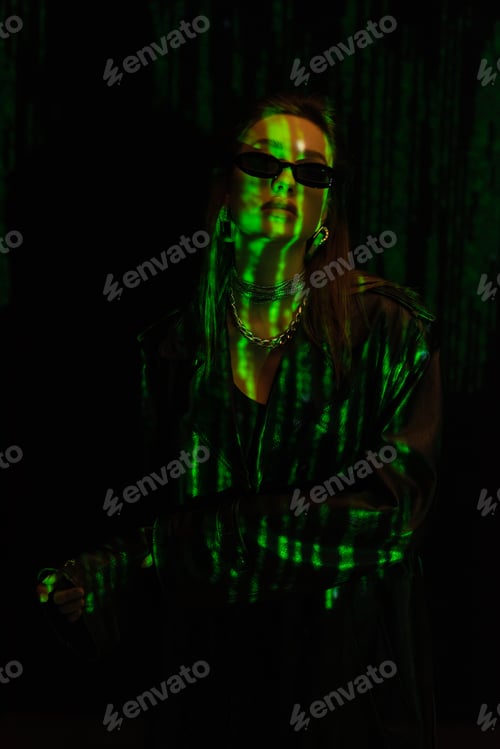 Preview: fashionable woman in sunglasses and silver accessories posing in green neon light isolated on black