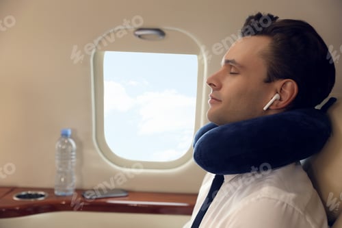 Preview: Young man with travel pillow resting while listening to music in airplane during flight