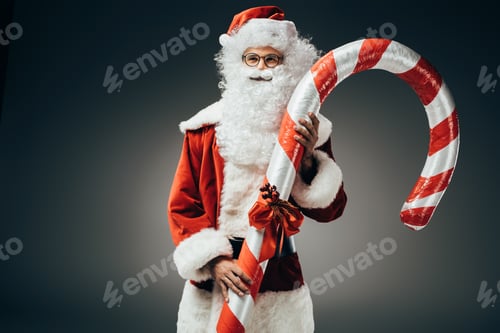 Preview: dreamy santa claus in costume standing with big striped christmas stick isolated on grey background
