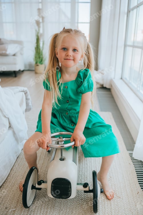 Preview: Smiling young girl in a vibrant green dress riding toy pedal car, enjoying playtime in sunlit room