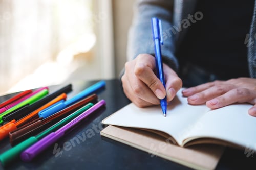 Preview: Closeup image of a woman writing on a blank notebook with colored pens on the table