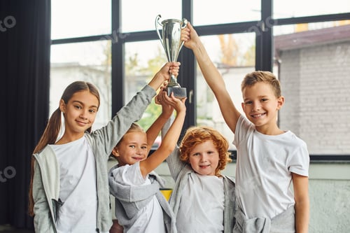 Preview: cheering preadolescent boys and girls in sportswear raising trophy above heads, child sport
