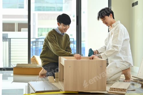 Preview: Cheerful young gay male couple using electric drill tool to assembling furniture in their new home