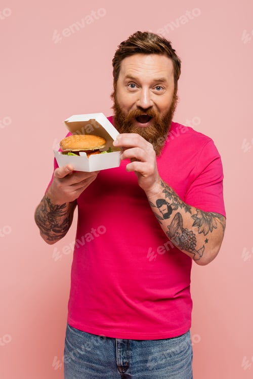 Preview: amazed man in magenta t-shirt showing carton box with tasty burger isolated on pink