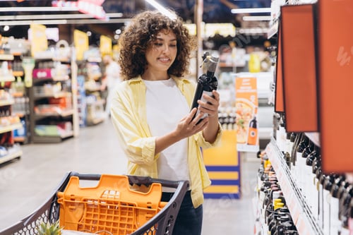 Preview: Woman Choosing Wine in Supermarket Reading Label with Shopping Cart