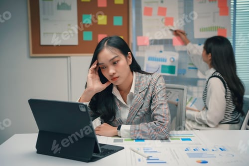 Preview: Stressed asian businesswoman looking at tablet with headache working in office meeting room