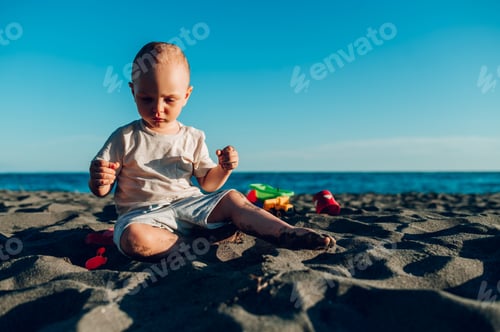 Preview: Caucasian little boy sitting in the sandy beach surrounded with toys.