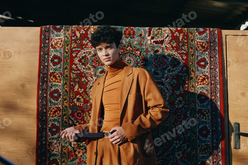 Preview: Young man in stylish brown outfit holds a gun in front of a colorful patterned tapestry
