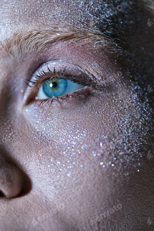 Preview: close up of young woman with blue eye and shiny sparkles on her face looking away, highlighter