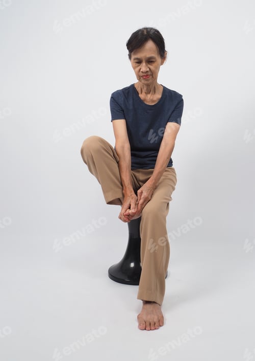 Preview: The senior or older woman grabs a foot and sits on a chair on white background.