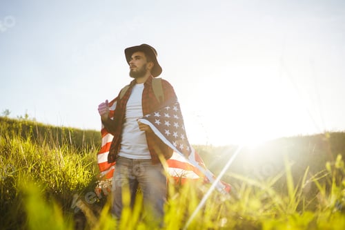 Preview: 4th of July. American Flag. Traveler with flag of America. Man in hat, backpack, a shirt and jeans.
