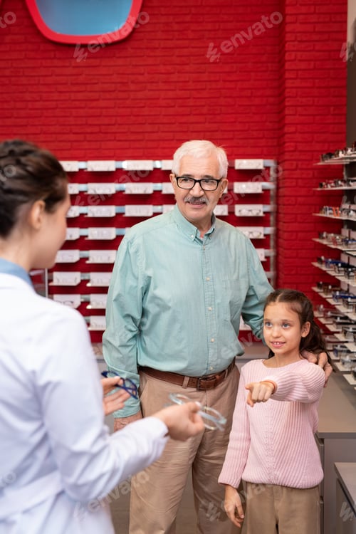 Preview: Adorable girl pointing at one of two pairs of eyeglasses held by ophthalmologist