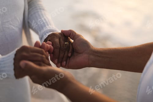 Preview: Midsection of newlywed multiracial senior couple holding hands at beach during sunset