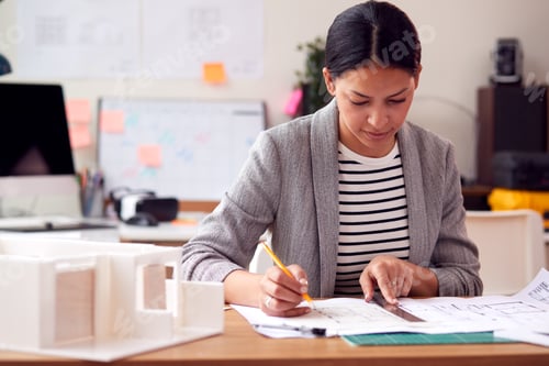 Preview: Female Architect Working In Office With Model On Desk Studying Plans For New Building