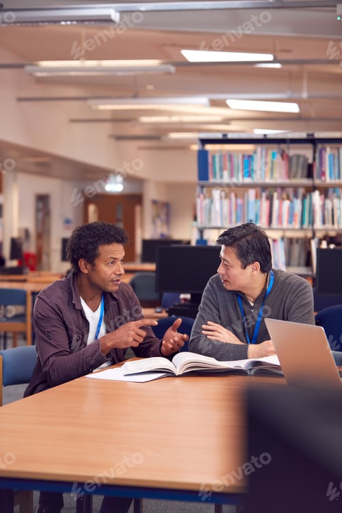 Preview: Teacher With Mature Male Adult Student Sitting At Table Working In College Library