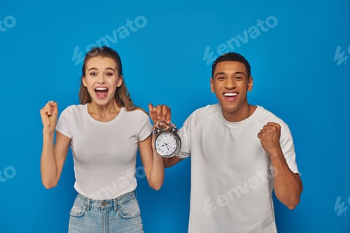 Preview: excited interracial couple with open mouth holding alarm clock on blue background, face expression