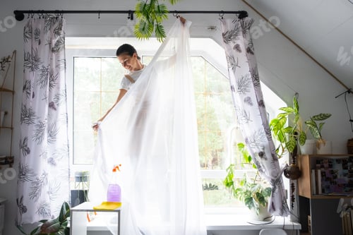 Preview: Woman in apron hangs transparent tulle curtains on large attic windows in the house inside
