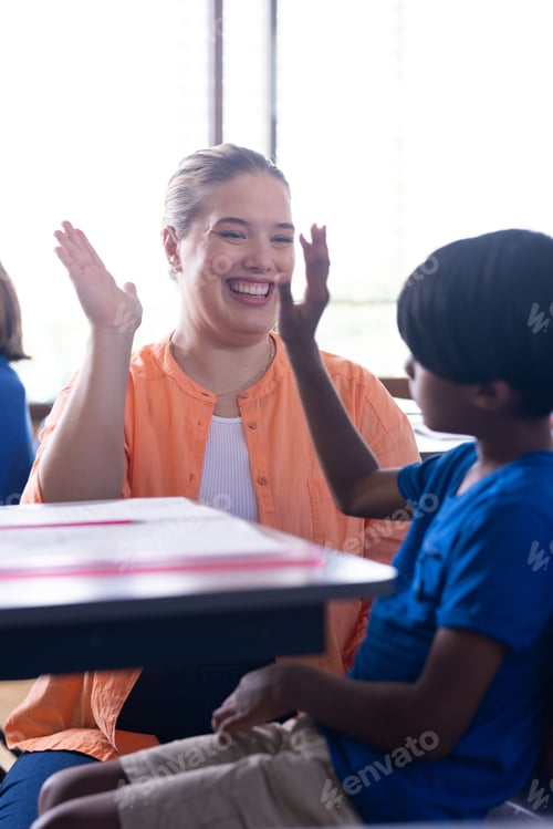 Preview: In school, female teacher and boy high-fiving while sitting at desk, smiling