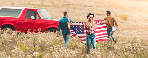 Preview: group of young friends with united states flag in flower field during road trip