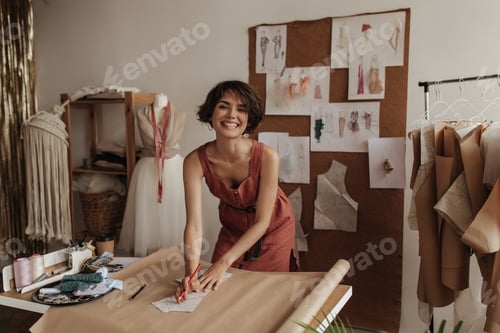Preview: Cheerful young fashion designer in linen red dress smiles sincerely. Charming woman holds pencils a