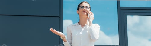 Preview: panoramic crop of happy businesswoman in glasses talking on smartphone near building