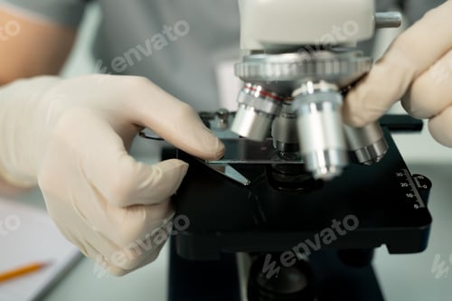 Preview: Gloved hand of scientist or clinician putting glass with chemical sample by lens