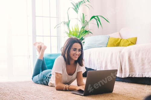 Preview: Happy young woman in eyeglasses typing using laptop while lying down on carpet in the living room