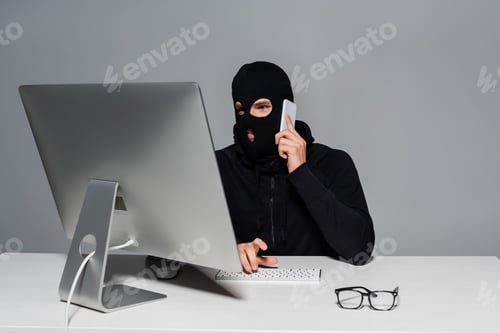 Preview: Hacker in balaclava talking on smartphone and using computer near eyeglasses isolated on grey